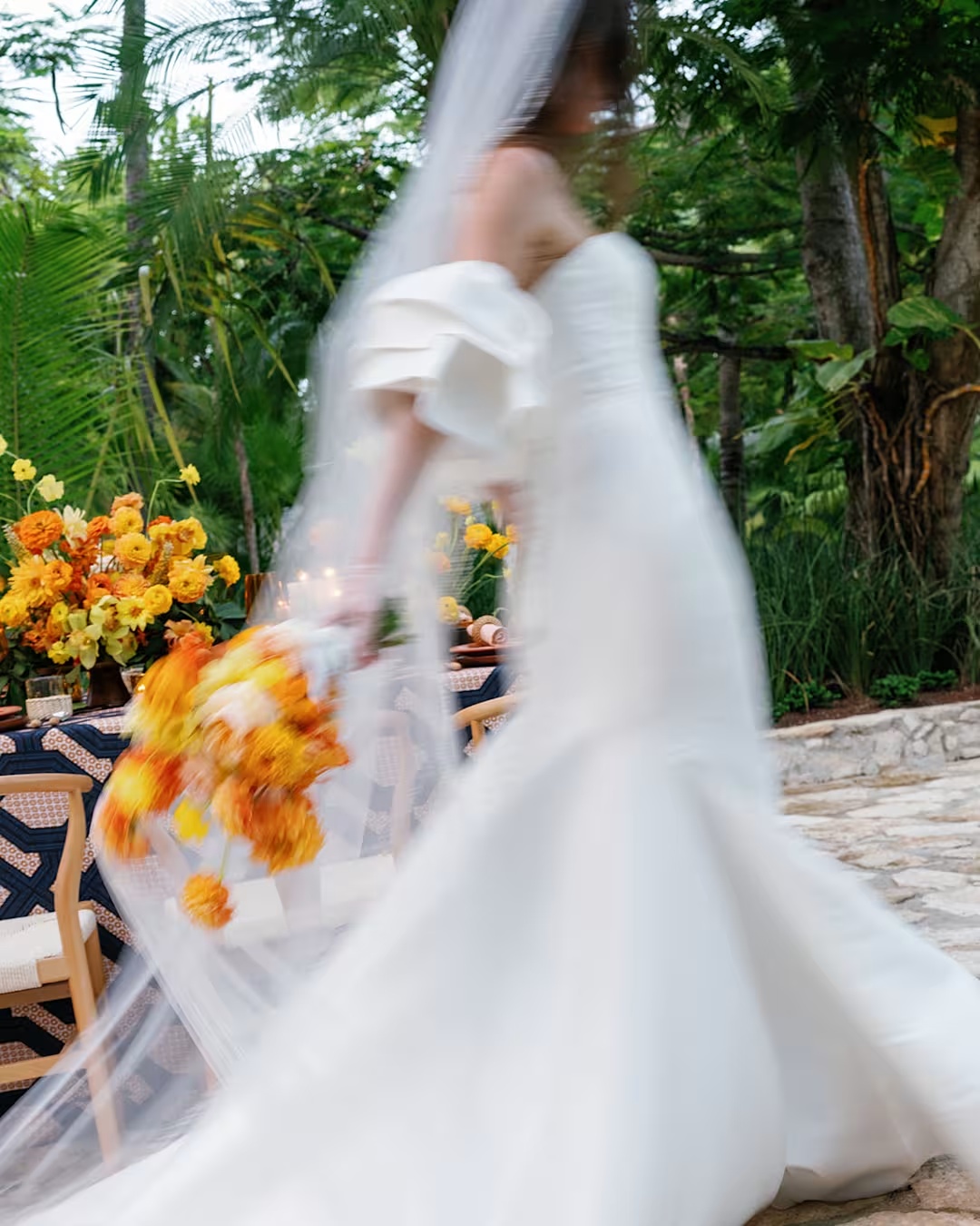 Bride in a flowing white gown holding an orange and yellow bouquet in a tropical garden